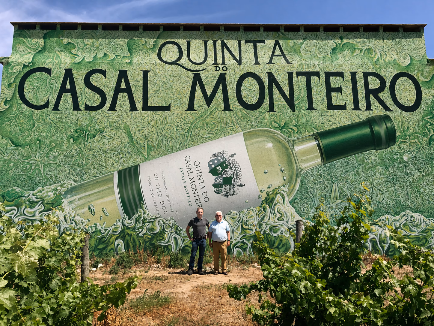 Miguel and Cirilo de Jesus standing in vineyard before Quinta do Casal Monteiro mural featuring oversized white wine bottle artwork